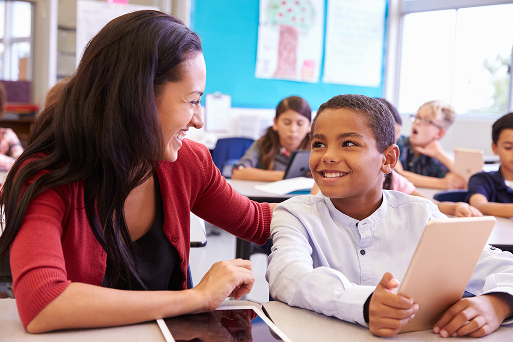 A female teacher looks over the shoulder of a young male student as he completes classwork on a tablet